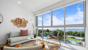 A modern living room with a large window, a sofa, a coffee table, and a wicker chair. - A Boho Chic Oceanfront Condo for 6. (Noord)
