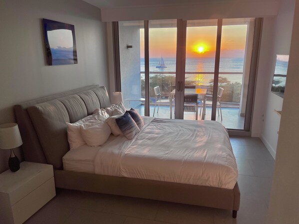 A bedroom with a bed, nightstand, and a view of the sunset over the ocean. - Imagine yourself in this Tropical Ocean-view Condo (Noord)