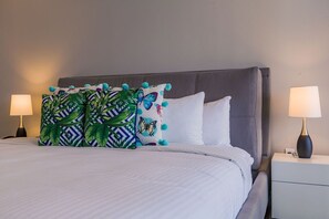 A neatly made bed with white linens, a grey upholstered headboard, and two bedside lamps on white nightstands. - Imagine yourself in this Tropical Ocean-view Condo (Noord)