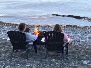 Two people seated at a table with a fire between them, on a rocky shore with water in the background.