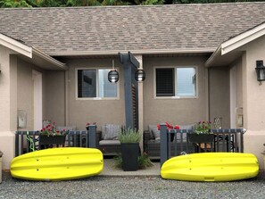 Un bâtiment avec un toit gris, deux fenêtres avec volets, un banc gris et deux kayaks jaunes devant.