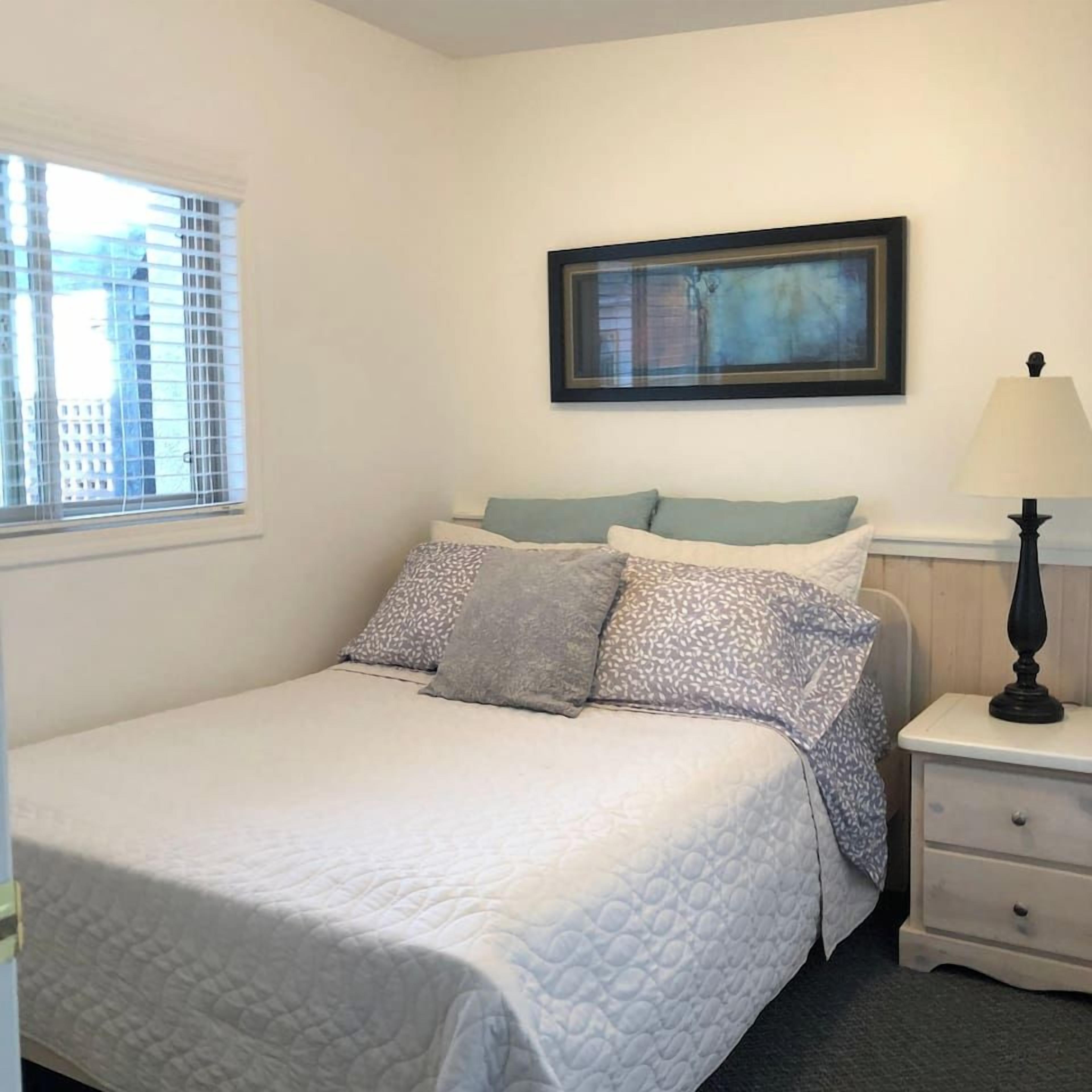 A bedroom with a bed, a nightstand, a lamp, a window with blinds, and a framed picture on the wall.