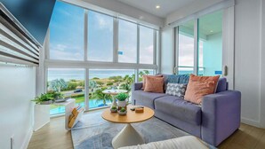 A modern living room with a large window, a sofa, a coffee table, and a view of the sea. - Sunsets are this Oceanview condo's best accessory! (Noord)