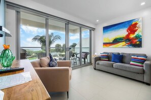 A modern living room with a large sliding glass door leading to a balcony, a grey sofa with colorful cushions, and a painting on the wall.