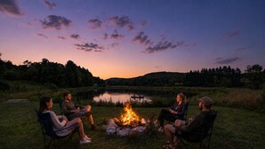 A group of people seated around a campfire in an outdoor setting with a lake and hills in the background. - 1/2 mile to EVL, Lake, HOT TUB, Pets welcome (Ellicottville)
