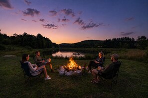 A group of people seated around a campfire in an outdoor setting with a lake and hills in the background. - 1/2 mile to EVL, Lake, HOT TUB, Pets welcome (Ellicottville)