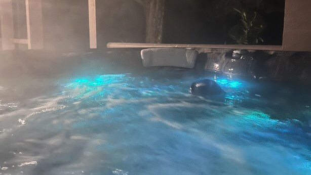 Ein Whirlpool mit blauer Beleuchtung, umgeben von einer Holzterrasse und einem überdachten Bereich.