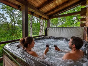 Un jacuzzi con dos personas sosteniendo copas de vino, rodeado de vigas de madera y vistas al follaje.