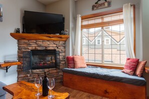 A cozy living room with a stone fireplace, a flat-screen TV mounted above it, a comfortable seating area with cushions, and a large window with blinds. - BigWhite Ski In/Out Chalet w Private HotTub & Garage (Beaverdell)