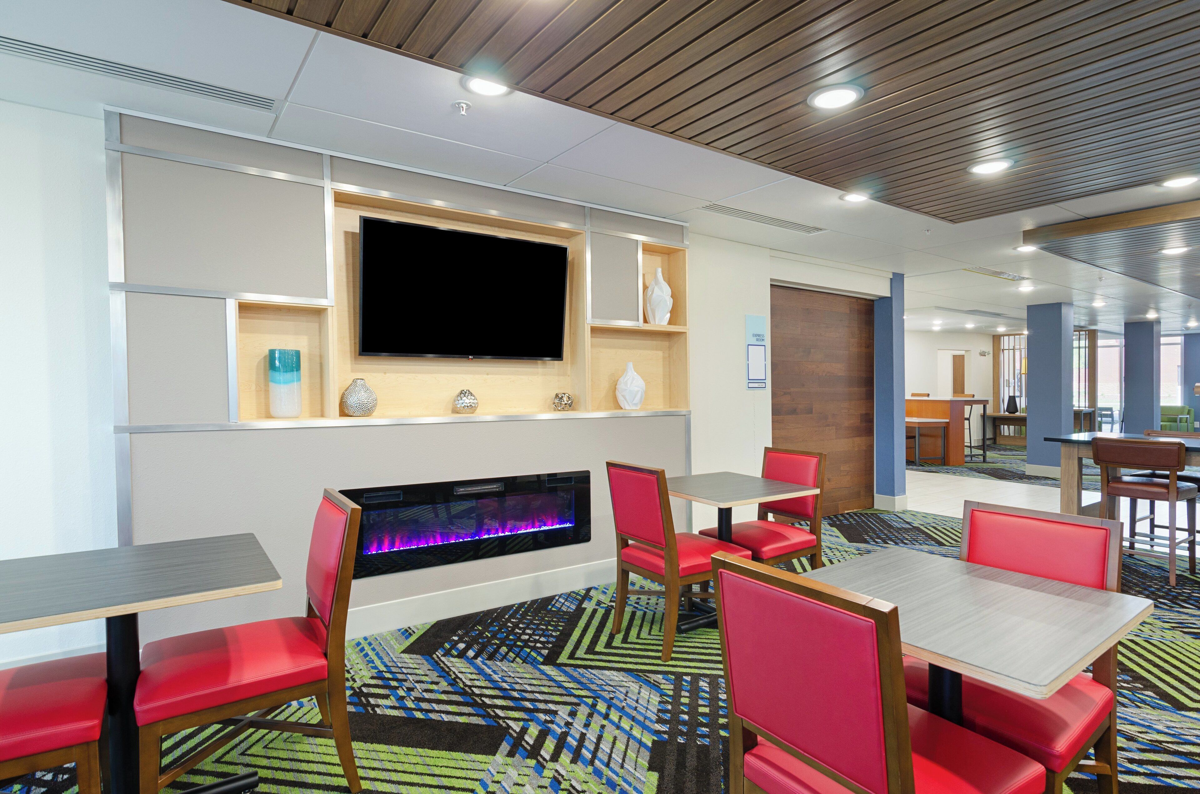 A hotel lobby with a flat-screen TV mounted on the wall, a fireplace with a colorful light display, red upholstered chairs and tables, and a patterned carpet.