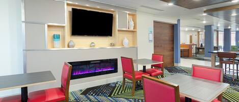 Un hall d’hôtel doté d’un téléviseur à écran plat fixé au mur, d’une cheminée avec un éclairage multicolore, de fauteuils et de tables recouverts d’un tissu rouge, et d’un tapis à motifs.