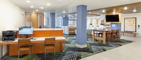 A modern hotel lobby with a reception desk, computer workstations, a dining area, and a TV screen.