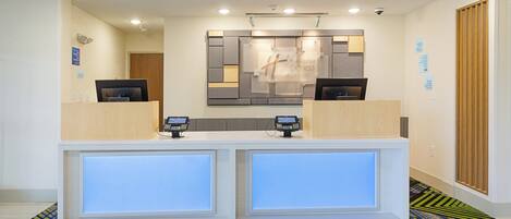 A hotel reception desk with two computer monitors, a digital display, and a "Thank You for Your Help" sign on the floor.