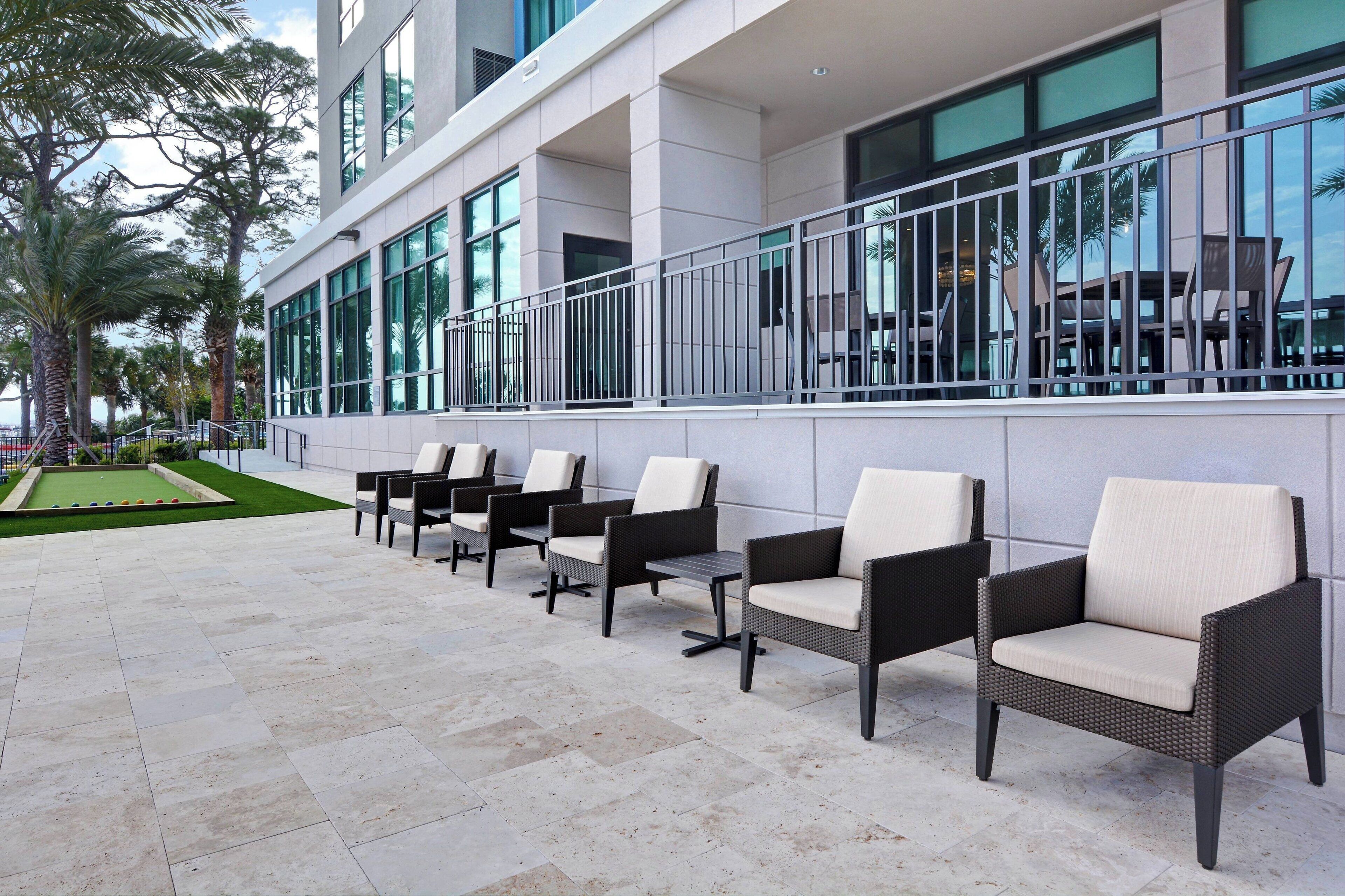 A row of outdoor wicker chairs with cushioned seats and backs, aligned against a building with large windows and a metal railing.