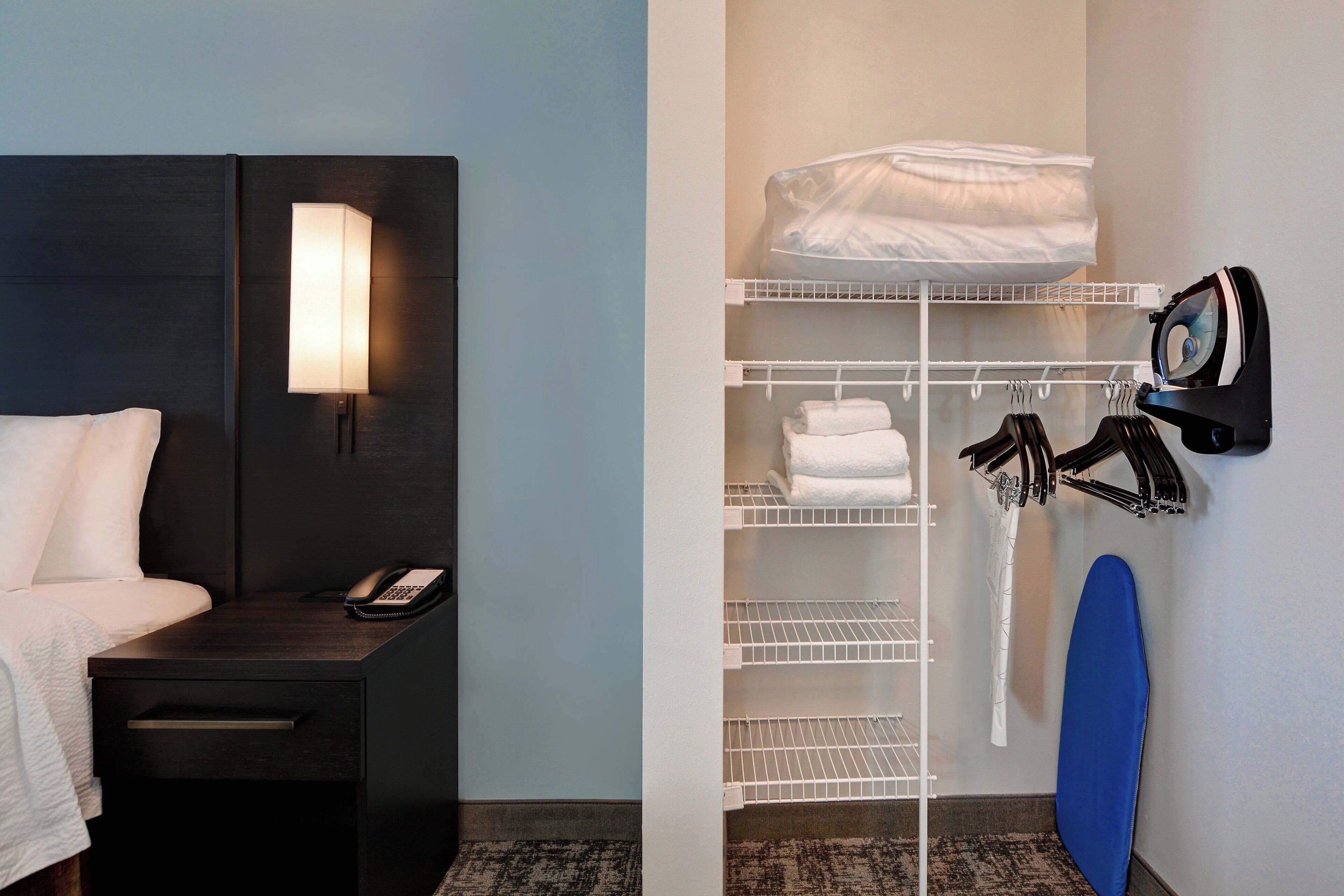 A hotel room with a bed, bedside table with a phone, a wall-mounted lamp, a closet with shelves, hanging clothes, and a blue mat.