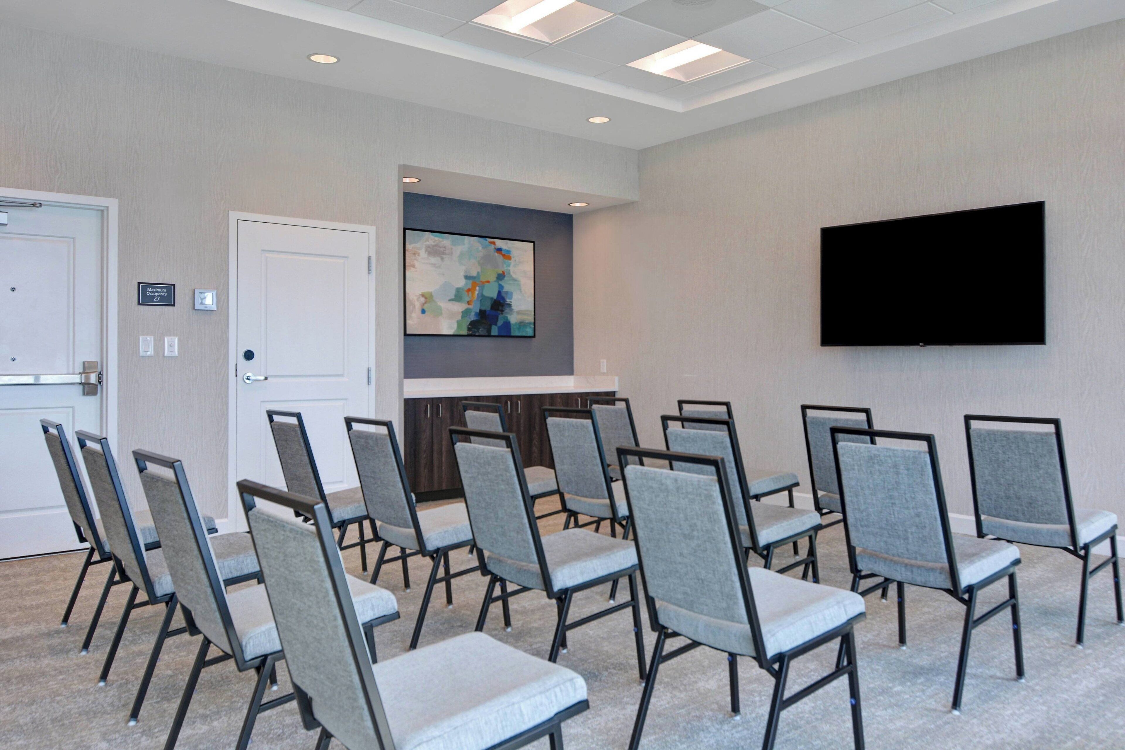A conference room with a flat-screen TV, grey cushioned chairs, a white door, and a painting on the wall.
