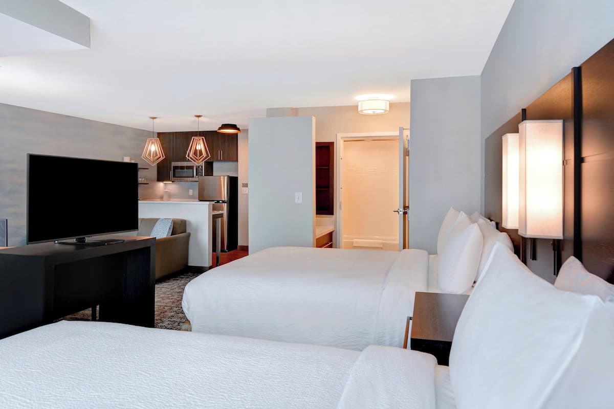 Ein Hotelzimmer mit zwei Betten, einem Fernseher, einem Schreibtisch, einer Mikrowelle und einer Küchenzeile.