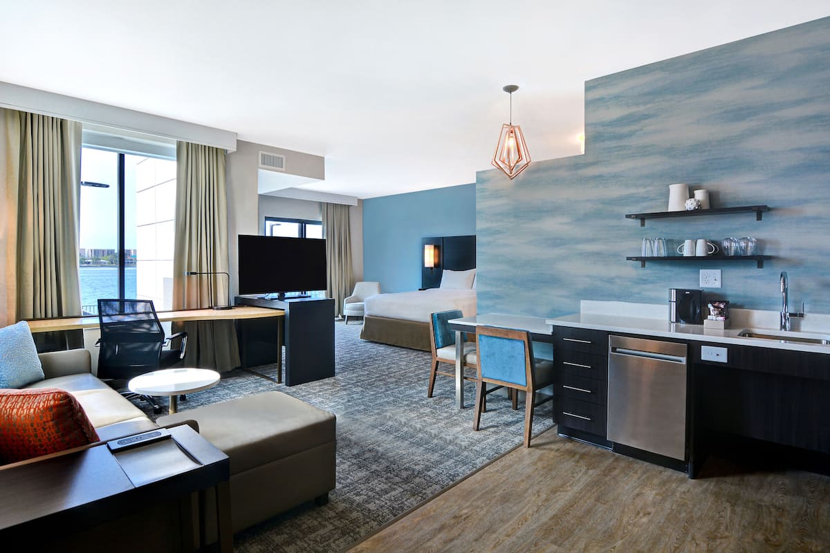 Ein modernes Hotelzimmer mit Wohnbereich, einem Esstisch, einer Couch, einem Fernseher, einer Küchenzeile und einem großen Fenster mit Vorhängen.