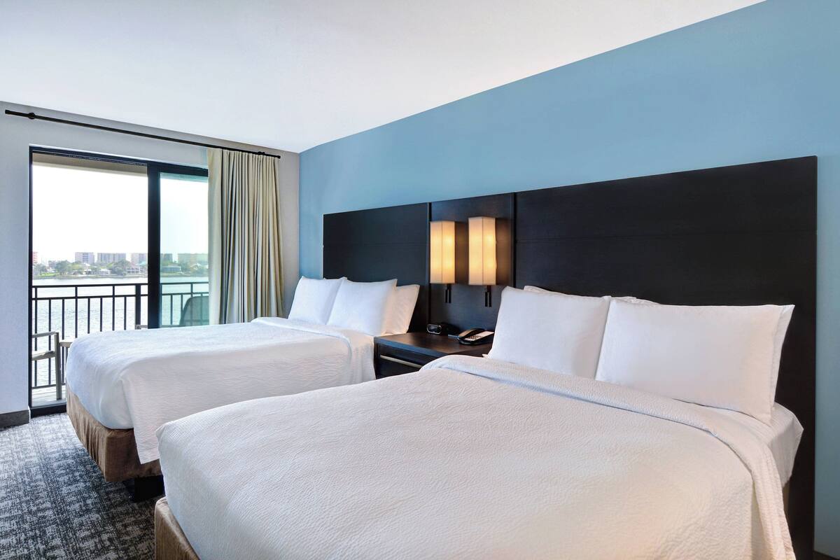 Ein Hotelzimmer mit zwei Betten, einem großen Fenster mit Vorhängen, einem Nachttisch mit Lampe und Blick auf die Stadt.