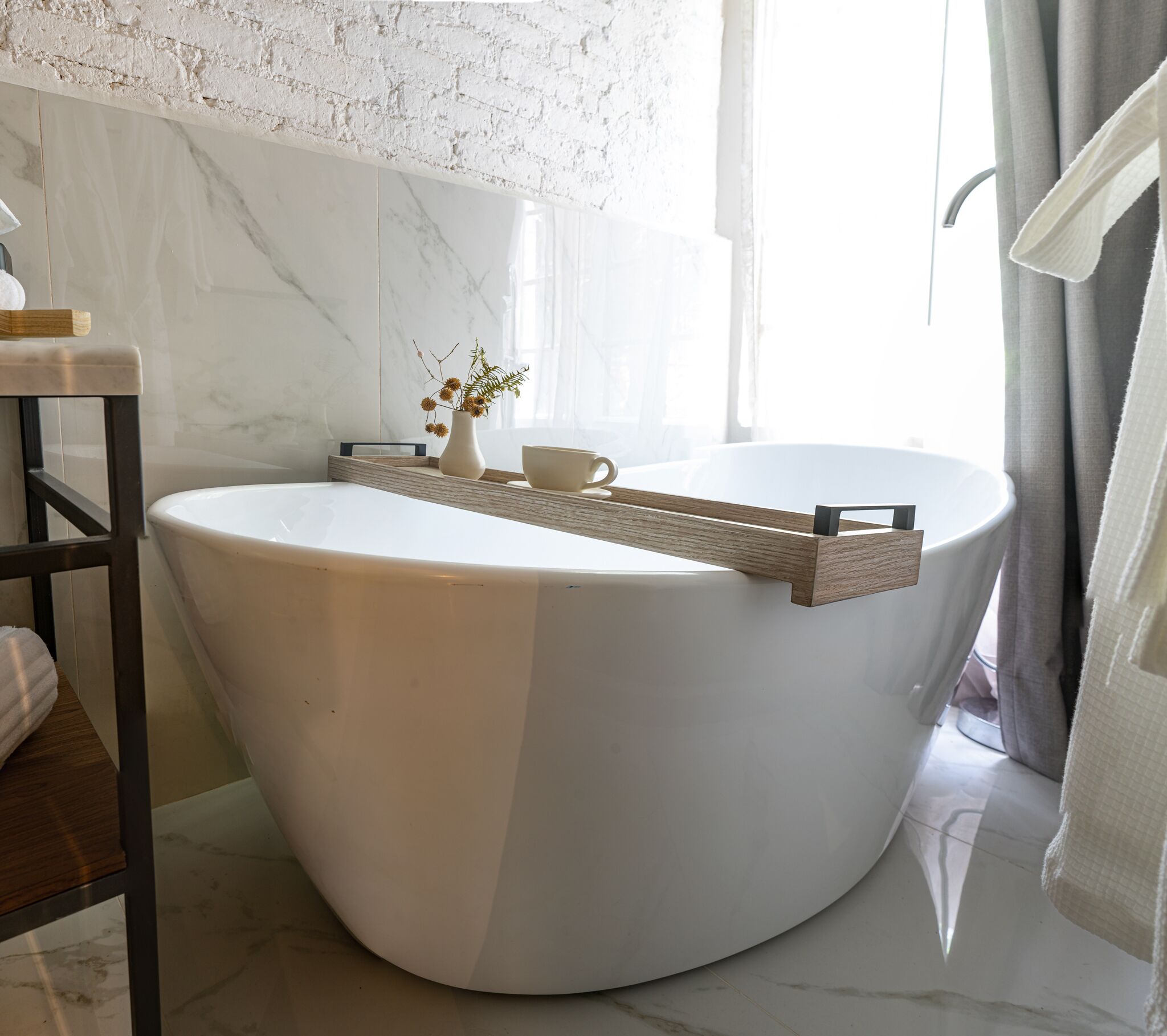 Een moderne badkamer met een vrijstaand bad, een houten dienblad, een vaas met bloemen en een beker.