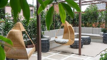 A covered outdoor seating area with wicker chairs and a cushioned bench, surrounded by potted plants and a pergola structure.