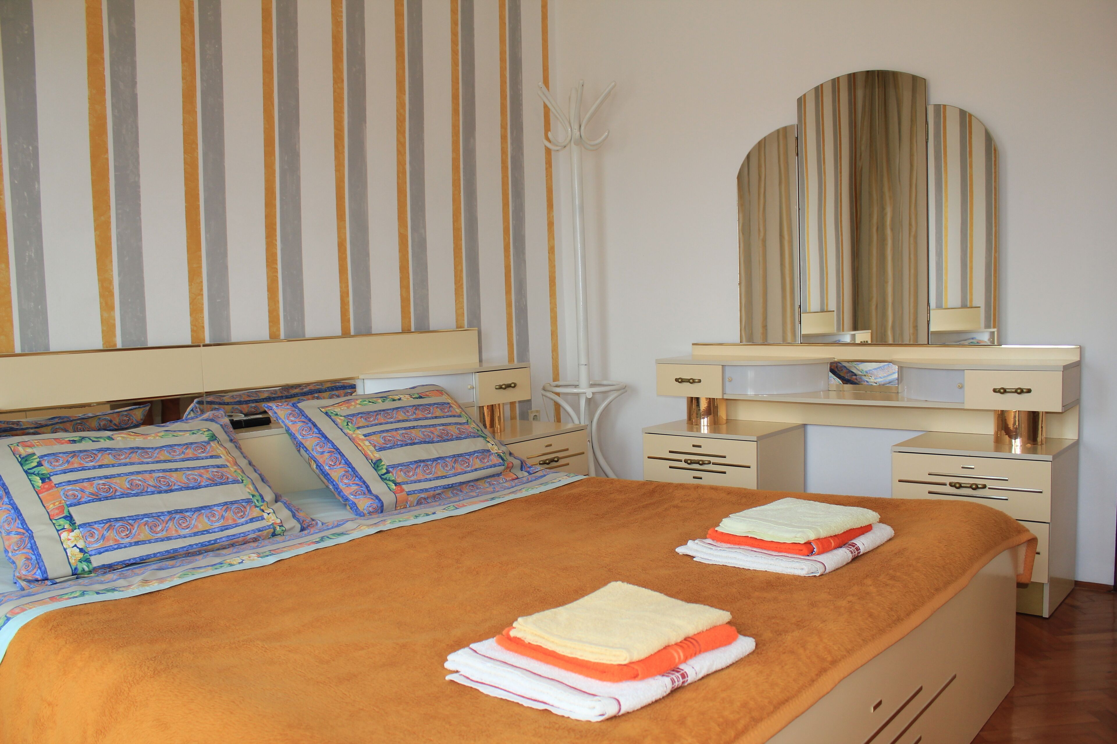 A neatly made bed with a striped headboard, a bedside table with a lamp, and a large mirror above a dresser.