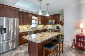 Una cocina moderna con gabinetes de madera, electrodomésticos de acero inoxidable, una isla de granito con taburetes y luminarias colgantes.