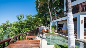 A modern outdoor pool area with a wooden deck, lounge chairs, and a white building with a balcony.