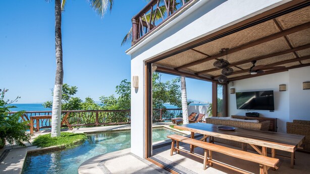 A poolside area with a wooden table and benches, a television, and a view of the ocean.
