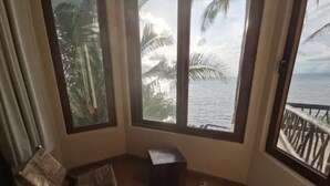 A corner room with two wicker chairs, a small table, and large windows offering a view of the ocean and palm trees.
