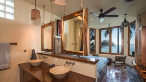 A bathroom with two vessel sinks, a large mirror, a ceiling fan, and a view of palm trees through large windows.