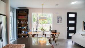 Una cucina moderna con una grande isola, tavolo da pranzo e sedie. Si trova una libreria piena di libri e bottiglie, un frigorifero e un divano bianco nella zona living.
