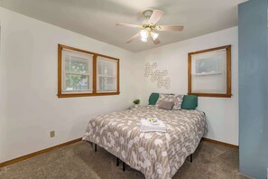 Un dormitorio con cama, dos ventanas con persianas, un ventilador de techo y un adorno en la pared.