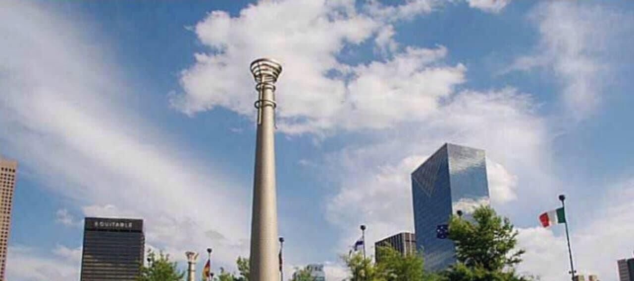 A tall, cylindrical structure with a pointed top, surrounded by modern buildings and flags.