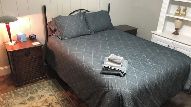 Ein ordentlich bezogenes Bett mit einem gemusterten Bettbezug und Kissen, ein Nachttisch mit Lampe und Uhr sowie ein kleiner Beistelltisch mit Schublade.