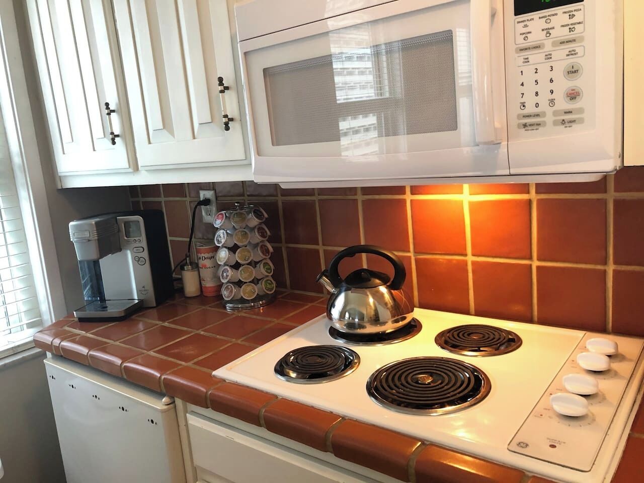 A kitchen with a microwave, stove, kettle, coffee maker, and a rack of bottles.