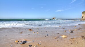 A beach with scattered shells and rocks, a rocky outcrop, and waves crashing onto the shore.