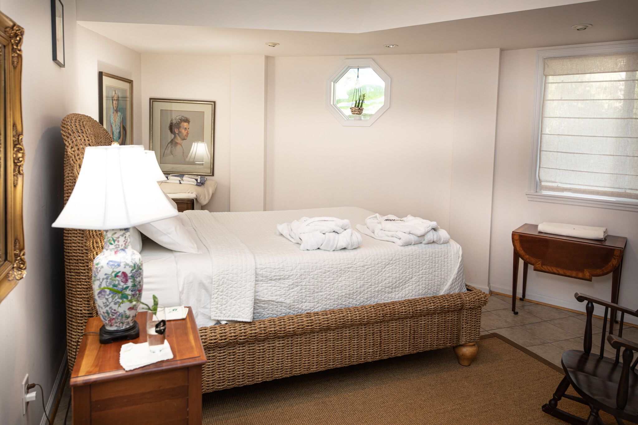 Un dormitorio ordenado con cama, lámpara de noche, cesta de mimbre y una mesita con lámpara.