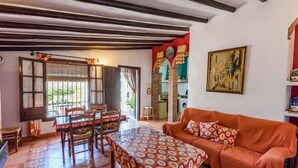 A cozy living room with a dining area, featuring a patterned sofa, wooden dining table, and a view of the kitchen through an open doorway. - El Ranchito de Martín Álora by Ruralidays (Málaga)