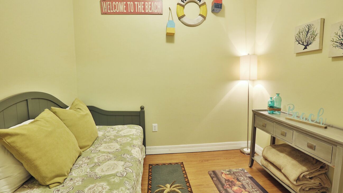 Un dormitorio con una cama, una mesita de noche, una lámpara y una pared con un cartel que dice 'Welcome to the Beach'.