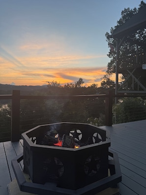 A deck with a fire pit, overlooking a body of water and a sunset.