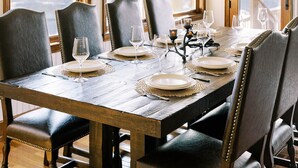 A dining room with a wooden table set for six, surrounded by leather chairs, with a view of mountains through large windows.