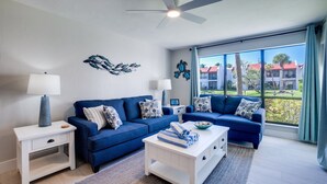 A modern living room with a blue sofa set, white side tables, and a ceiling fan. - Miracle's Runaway Bay 117 by SeaBreeze Vacation (Bradenton Beach)