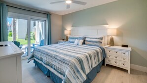 A bedroom with a bed, nightstands, a ceiling fan, and a view of the outdoors. - Miracle's Runaway Bay 117 by SeaBreeze Vacation (Bradenton Beach)