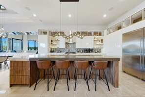 A modern kitchen with a large island, bar stools, a stainless steel refrigerator, and a decorative light fixture. - Reunion Mansion with Infinity Pool & Game Room (Kissimmee)