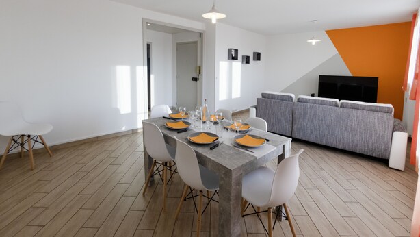 Un comedor moderno con una mesa rectangular dispuesta para cuatro personas, rodeada de sillas blancas con patas de madera. Se ve un sofá gris y un televisor al fondo.