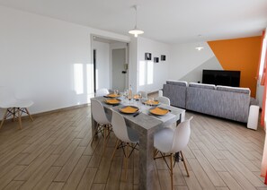 Una zona de menjador moderna amb una taula rectangular preparada per a quatre, envoltada per caires blancs amb potes de fusta. Es veu un sofà gris i una televisió al fons.