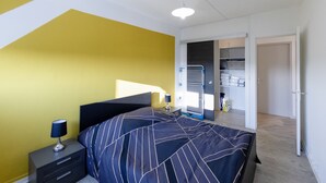 Un dormitorio con cama, mesitas de noche, lámpara, armario y una puerta que da a otra habitación.