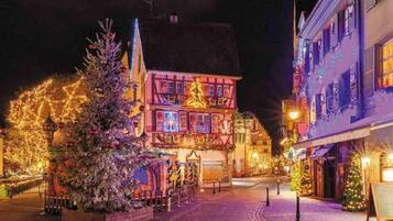 A traditional European town square at night, featuring a brightly lit building with a Christmas tree in the foreground.