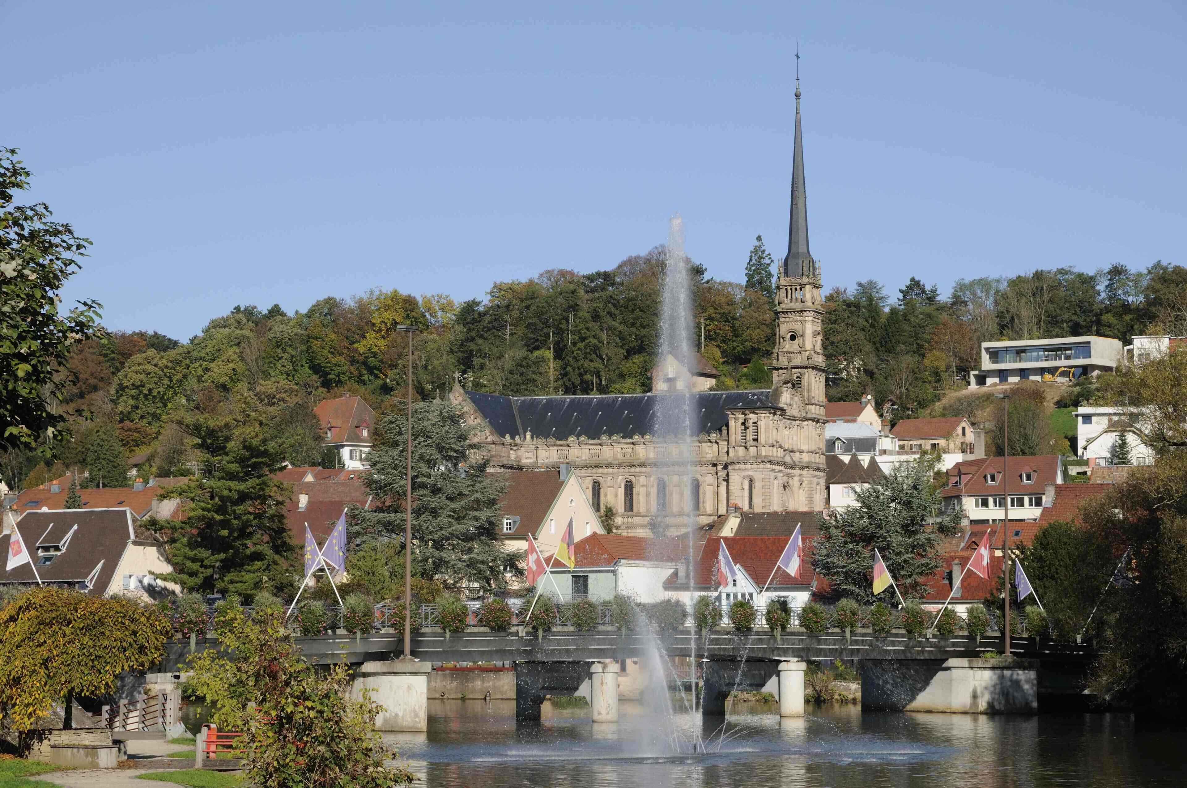Ein historisches Gebäude mit einem hohen Turm, einem Brunnen im Vordergrund und Flaggen auf einer Brücke.